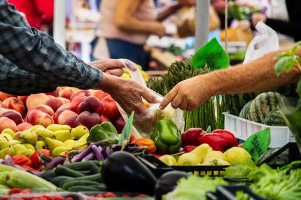 a buyer and a vendor exchange fresh vegetables at a busy farmers market. colorful produce including peppers, peaches, eggplants, asparagus, and other seasonal items fills the foreground.