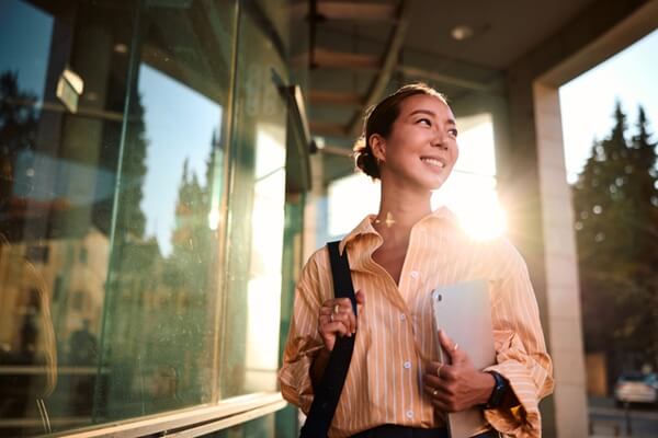 Young businesswoman walking confidently near an office building, holding a tablet and smiling while looking away, beautifully backlit by the warm glow of the setting sun