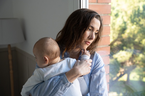 New mom holding her infant and looking out the window, overwhelmed by postpartum depression