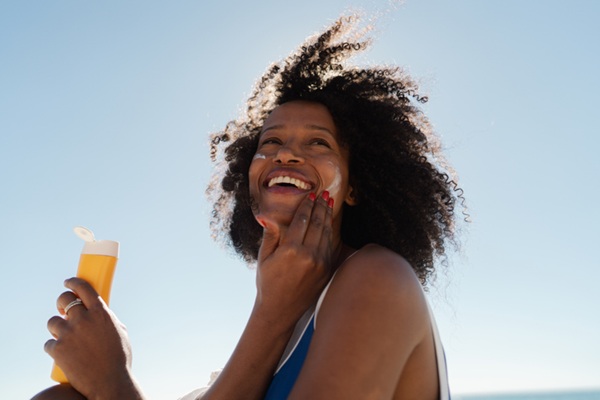 Photo of an African American woman applying sunscreen on her face while sunbathing at the beach, because darker complexion also needs protection.